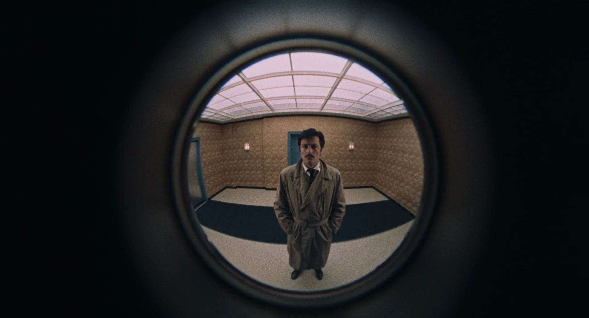 A man in a trench coat, seen through a door peephole, standing in an empty hallway